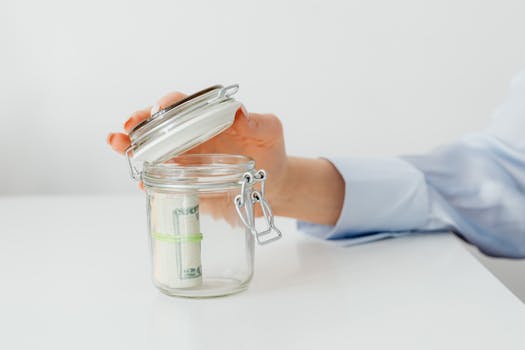 A hand places money in a glass jar on a white table, symbolizing savings.