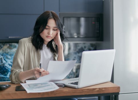 Focused young woman reviewing financial documents with a laptop in a home office setting.